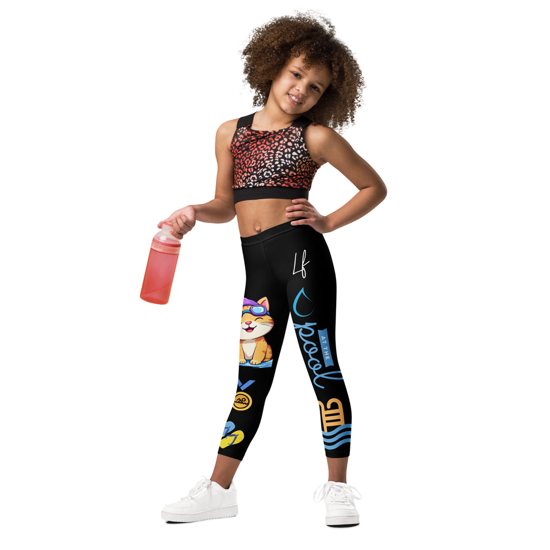Aqua Kitty Winner Girls Leggings, black background with a blue and yellow swimmer cat wearing a diving mask and swim cap on one leg, golden medal and pool slippers below, "at the pool" text with water drop, and pool water with ladder on the other leg, relaxed front pose on model