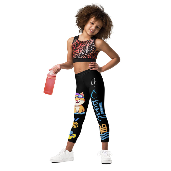 Aqua Kitty Winner Girls Leggings, black background with a blue and yellow swimmer cat wearing a diving mask and swim cap on one leg, golden medal and pool slippers below, "at the pool" text with water drop, and pool water with ladder on the other leg, relaxed front pose on model