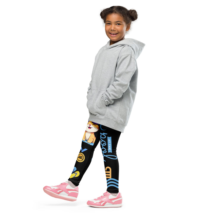 Aqua Kitty Winner Girls Leggings, black background with a blue and yellow swimmer cat wearing a diving mask and swim cap on one leg, golden medal and pool slippers below, "at the pool" text with water drop, and pool water with ladder on the other leg, side view with one leg lifted and extended on model