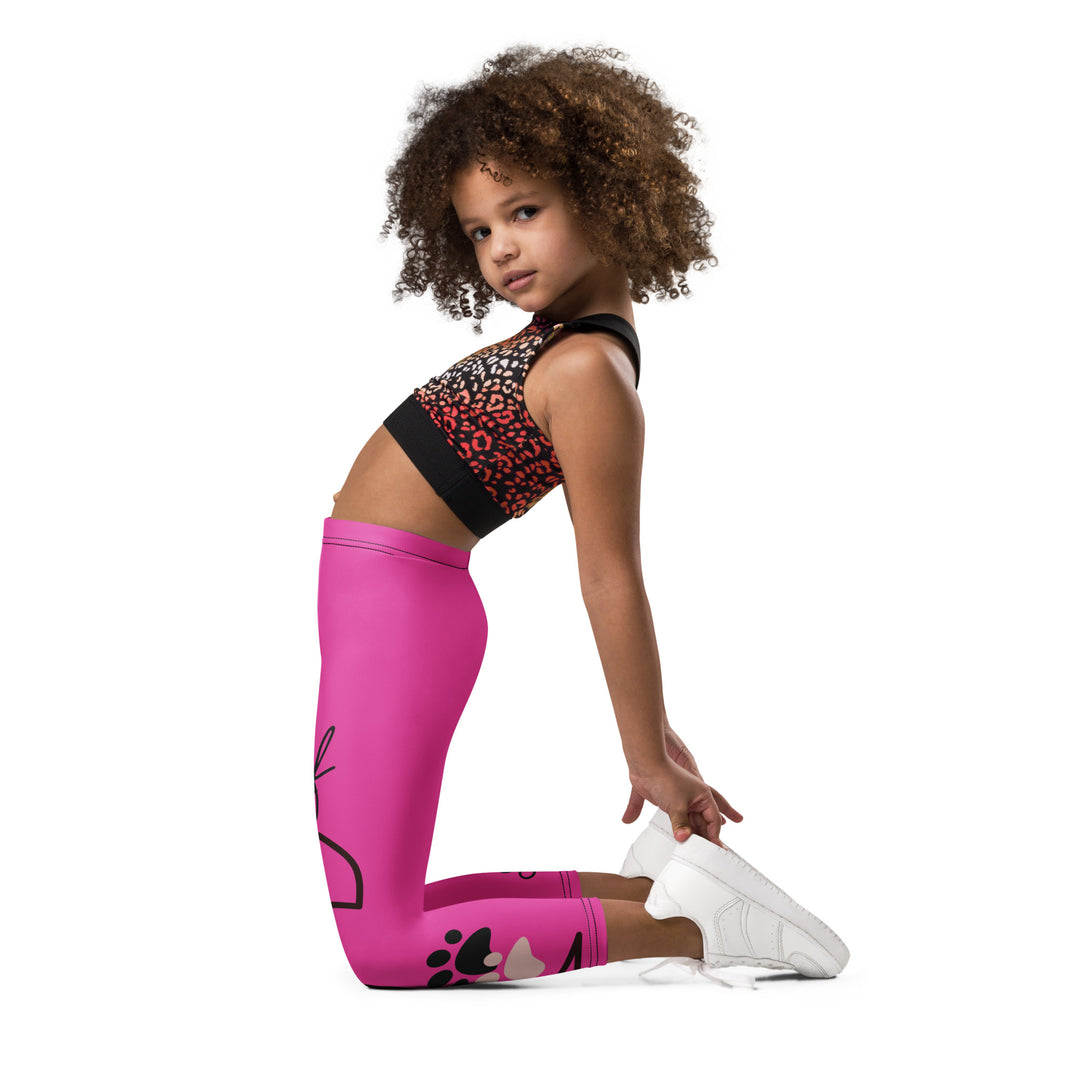  Cat Trace Girls Leggings, pink background with a yellow cat wearing a pink dress at the top of one leg, black paw prints and a black cat nose below, and a black line-drawn cat with black and cream paw prints on the other leg, left side view kneeling on model