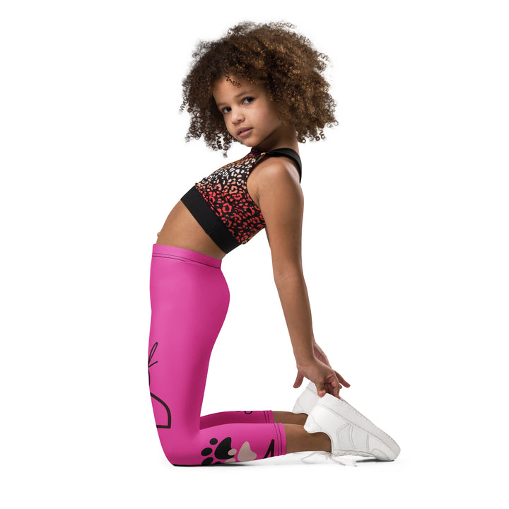 Cat Trace Girls Leggings, pink background with a yellow cat wearing a pink dress at the top of one leg, black paw prints and a black cat nose below, and a black line-drawn cat with black and cream paw prints on the other leg, left side view kneeling on model