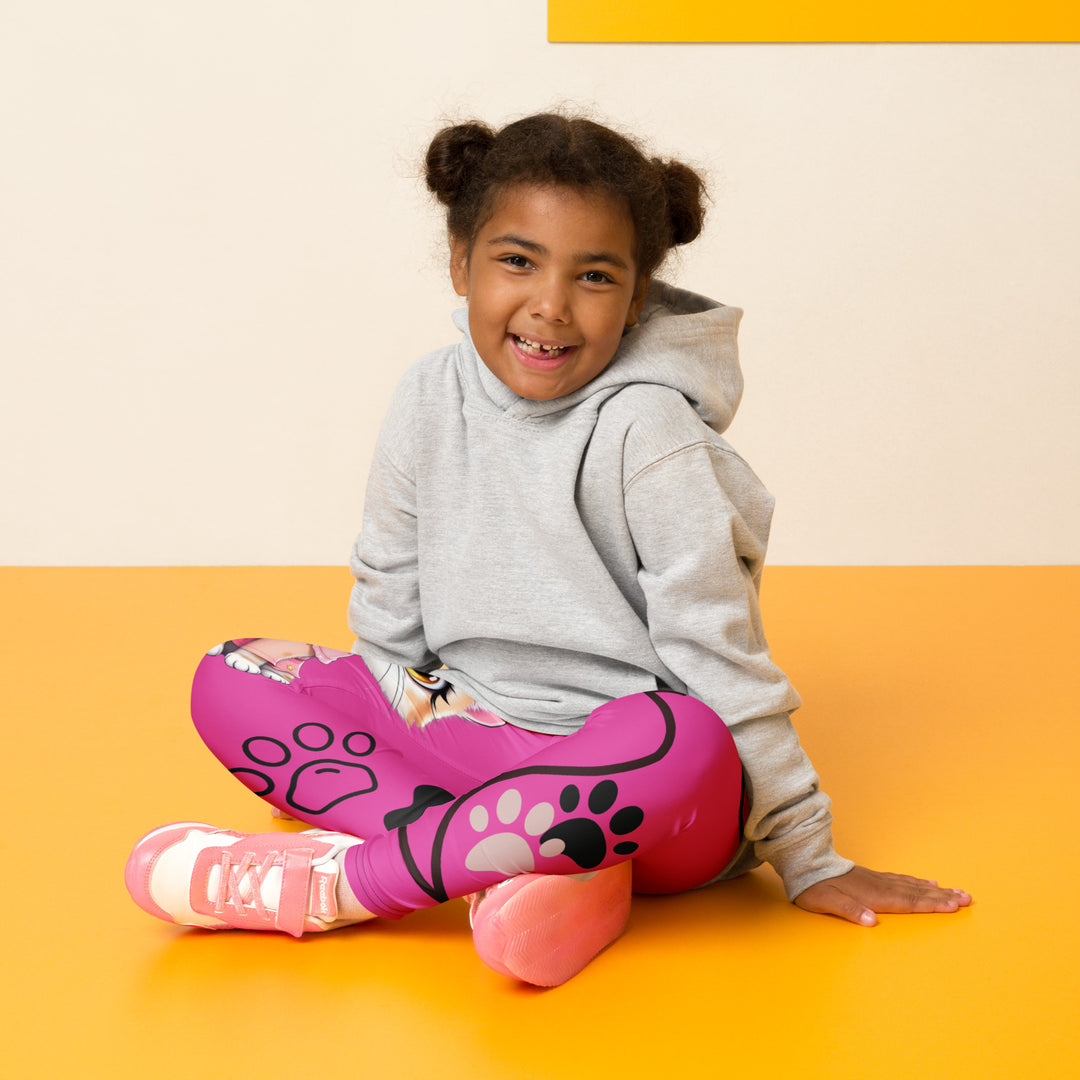  Cat Trace Girls Leggings, pink background with a yellow cat wearing a pink dress at the top of one leg, black paw prints and a black cat nose below, and a black line-drawn cat with black and cream paw prints on the other leg, seated pose on model