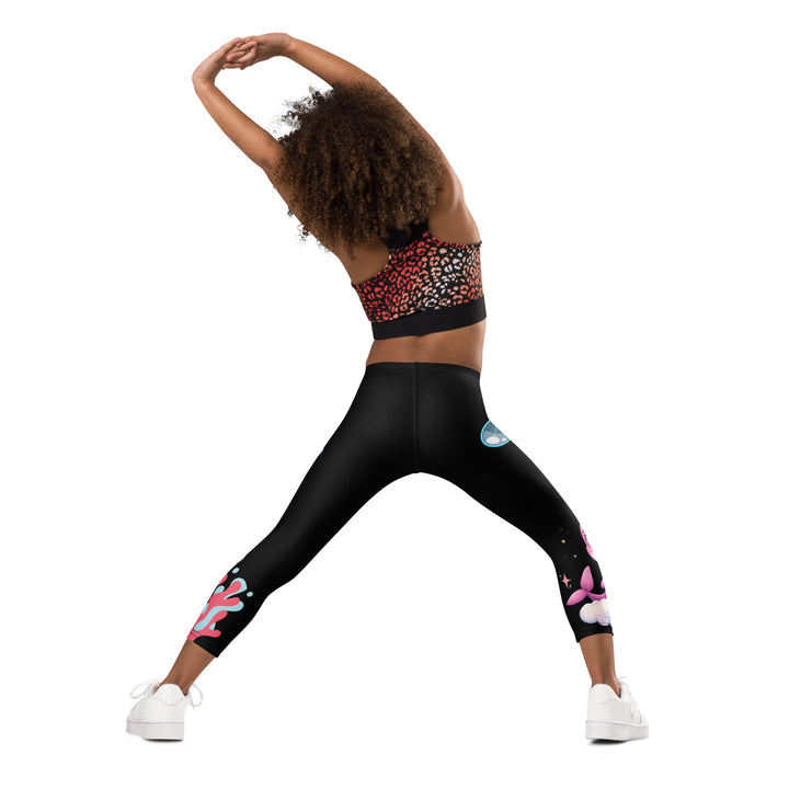  Coral Dolphin Girls Leggings, black background with a pink baby dolphin lying on a white cloud at the bottom of one leg and blue sea bubbles above, and a blue baby dolphin on the upper part of the other leg with pink and blue coral details at the bottom, back angle with relaxed pose on model
