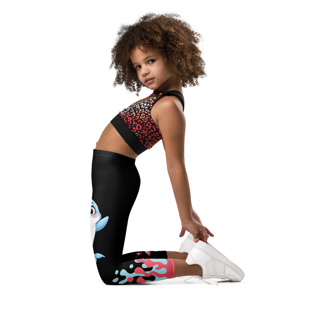 Coral Dolphin Girls Leggings, black background with a pink baby dolphin lying on a white cloud at the bottom of one leg and blue sea bubbles above, and a blue baby dolphin on the upper part of the other leg with pink and blue coral details at the bottom, left side view kneeling on model