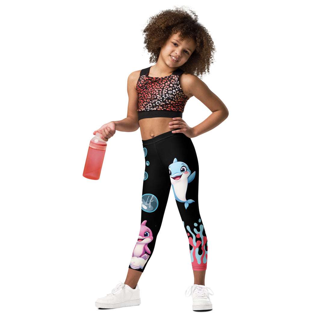 Coral Dolphin Girls Leggings, black background with a pink baby dolphin lying on a white cloud at the bottom of one leg and blue sea bubbles above, and a blue baby dolphin on the upper part of the other leg with pink and blue coral details at the bottom, relaxed front pose on model