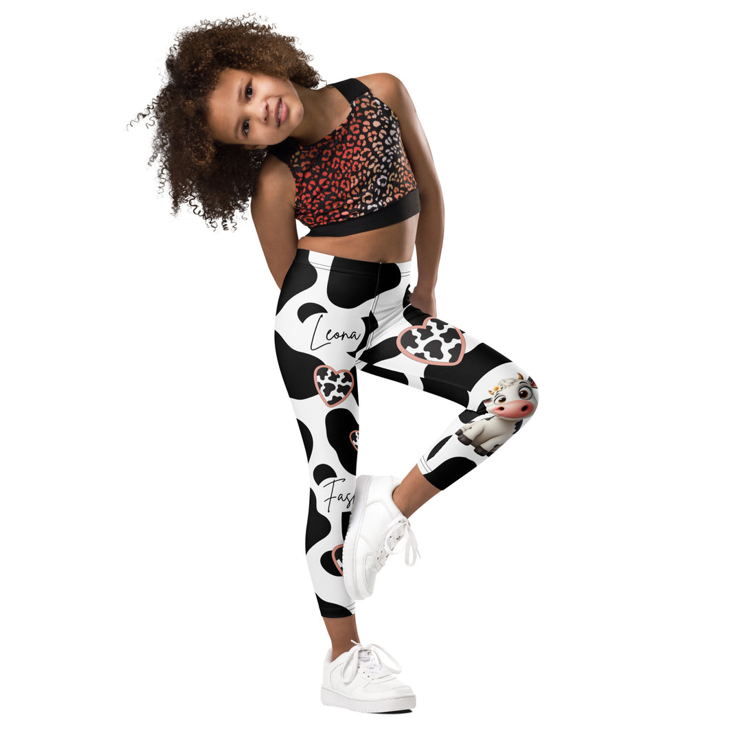 Cowlicious Love Girls Leggings, white background with black cow-like spots and a small black and white cow with a flower in its hair on one leg, front view with one leg lifted and bent on model-with a visible 'Leona Fashion' text on the design