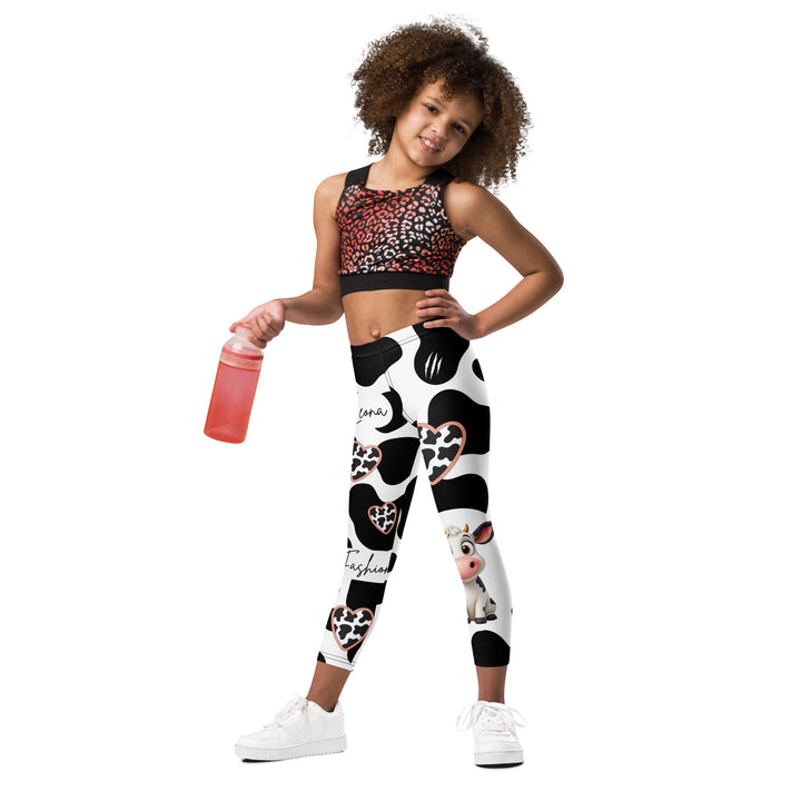 Cowlicious Love Girls Leggings, white background with black cow-like spots and a small black and white cow with a flower in its hair on one leg, relaxed front pose on model-with a visible 'Leona Fashion' text on the design