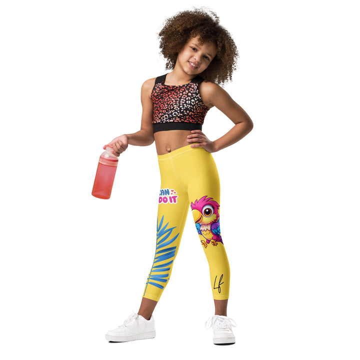 Fly Baby Parrot Girls Leggings, yellow background with a small pink baby parrot with spread wings on one leg, and a large blue tropical leaf with "I CAN DO IT" text in blue and pink letters above it on the other leg, relaxed front pose on model