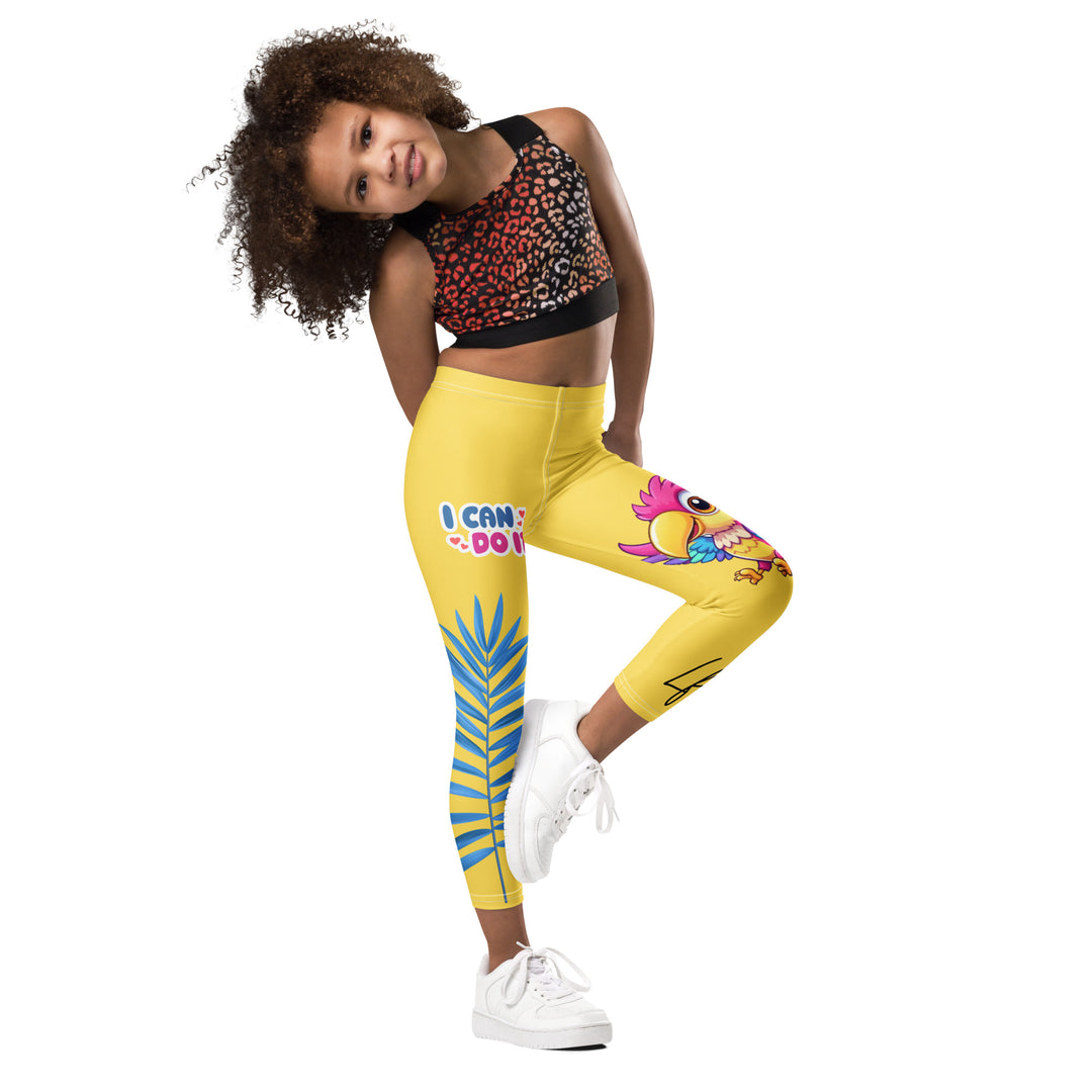  Fly Baby Parrot Girls Leggings, yellow background with a small pink baby parrot with spread wings on one leg, and a large blue tropical leaf with "I CAN DO IT" text in blue and pink letters above it on the other leg, side view with one leg lifted and bent on model