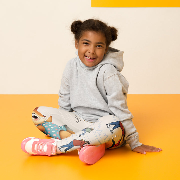 Frosty Twirl Squirrel Girls Leggings, beige background with a brown squirrel ice skating in a blue polka dot dress on one leg, ice trail lines, mountains, and small ice skaters along the other leg, in blue, brown, and orange tones, seated pose on model
