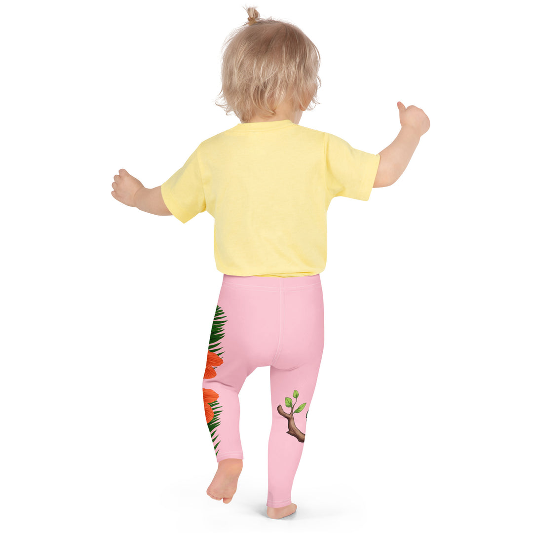 Jungle Parrot Girls Leggings, light pink background with a red, yellow, orange, and green parrot standing on a branch on one leg, and green jungle leaves with two large orange flowers running down the other leg, back pose on model