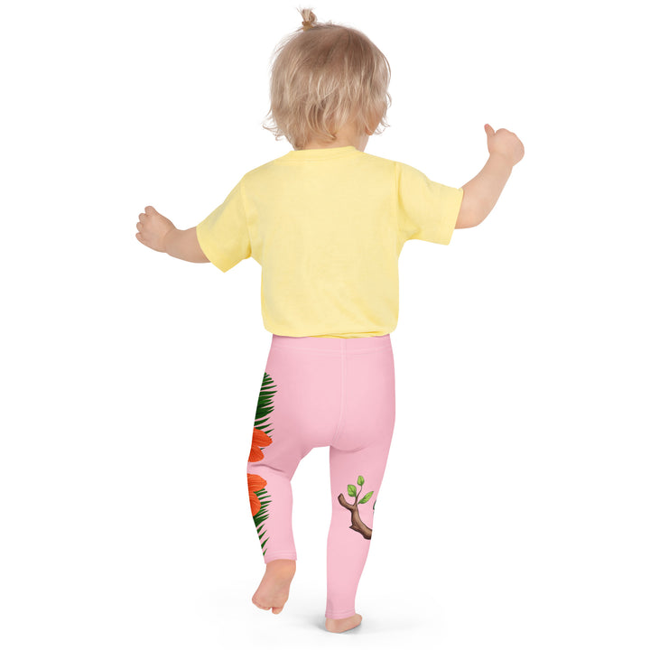 Jungle Parrot Girls Leggings, light pink background with a red, yellow, orange, and green parrot standing on a branch on one leg, and green jungle leaves with two large orange flowers running down the other leg, back pose on model
