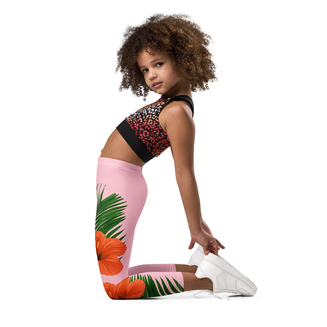  Jungle Parrot Girls Leggings, light pink background with a red, yellow, orange, and green parrot standing on a branch on one leg, and green jungle leaves with two large orange flowers running down the other leg, left side view kneeling on model