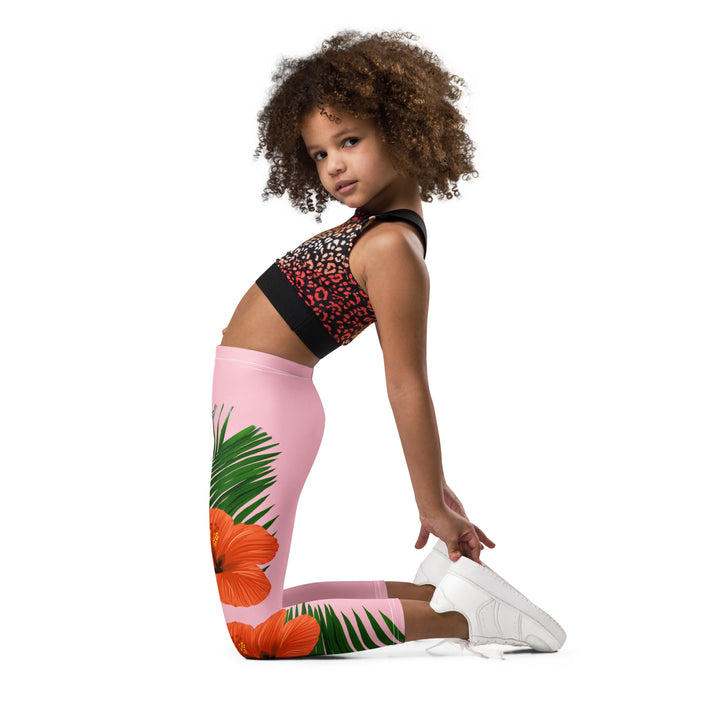  Jungle Parrot Girls Leggings, light pink background with a red, yellow, orange, and green parrot standing on a branch on one leg, and green jungle leaves with two large orange flowers running down the other leg, left side view kneeling on model