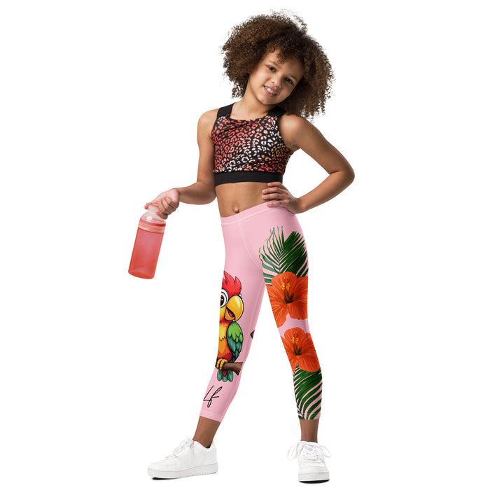Jungle Parrot Girls Leggings, light pink background with a red, yellow, orange, and green parrot standing on a branch on one leg, and green jungle leaves with two large orange flowers running down the other leg, relaxed front pose on model