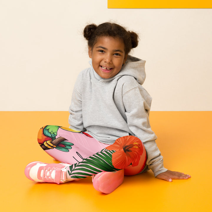 Jungle Parrot Girls Leggings, light pink background with a red, yellow, orange, and green parrot standing on a branch on one leg, and green jungle leaves with two large orange flowers running down the other leg, seated pose on model