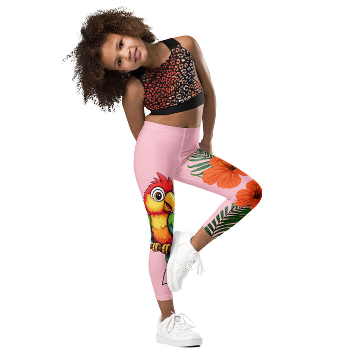 Jungle Parrot Girls Leggings, light pink background with a red, yellow, orange, and green parrot standing on a branch on one leg, and green jungle leaves with two large orange flowers running down the other leg, side view with one leg lifted and bent on model