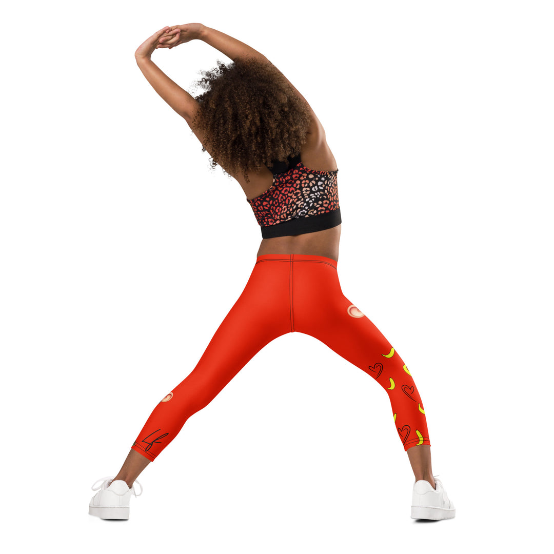 Love Monkey Girls Leggings, red background with a smiling female monkey with big eyes and a pink bow on the head at the top of one leg, yellow bananas with black drawn hearts below, and a winking male monkey wearing a hat sitting on a tree stump near the bottom of the other leg, with the word "LOVE" above, back angle with relaxed pose on model