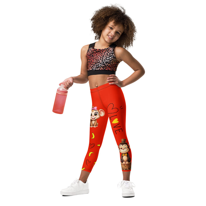Love Monkey Girls Leggings, red background with a smiling female monkey with big eyes and a pink bow on the head at the top of one leg, yellow bananas with black drawn hearts below, and a winking male monkey wearing a hat sitting on a tree stump near the bottom of the other leg, with the word "LOVE" above, relaxed front pose on model