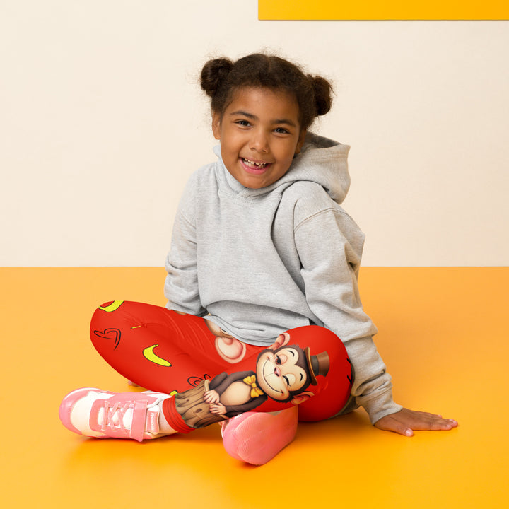 Love Monkey Girls Leggings, red background with a smiling female monkey with big eyes and a pink bow on the head at the top of one leg, yellow bananas with black drawn hearts below, and a winking male monkey wearing a hat sitting on a tree stump near the bottom of the other leg, with the word "LOVE" above, seated pose on model