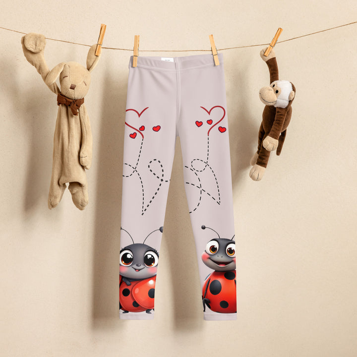 Lovebugs Girls Leggings, sand-colored background with a female ladybug with big eyes at the bottom of one leg and a male ladybug looking delighted on the other leg, with black swirling lines above them forming hearts on both sides, front view on hanger