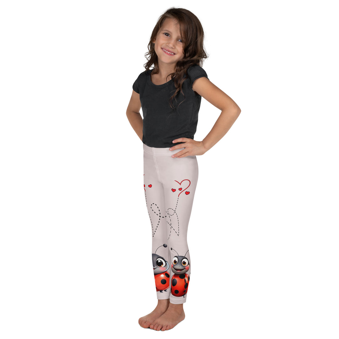Lovebugs Girls Leggings, sand-colored background with a female ladybug with big eyes at the bottom of one leg and a male ladybug looking delighted on the other leg, with black swirling lines above them forming hearts on both sides, left side view on model
