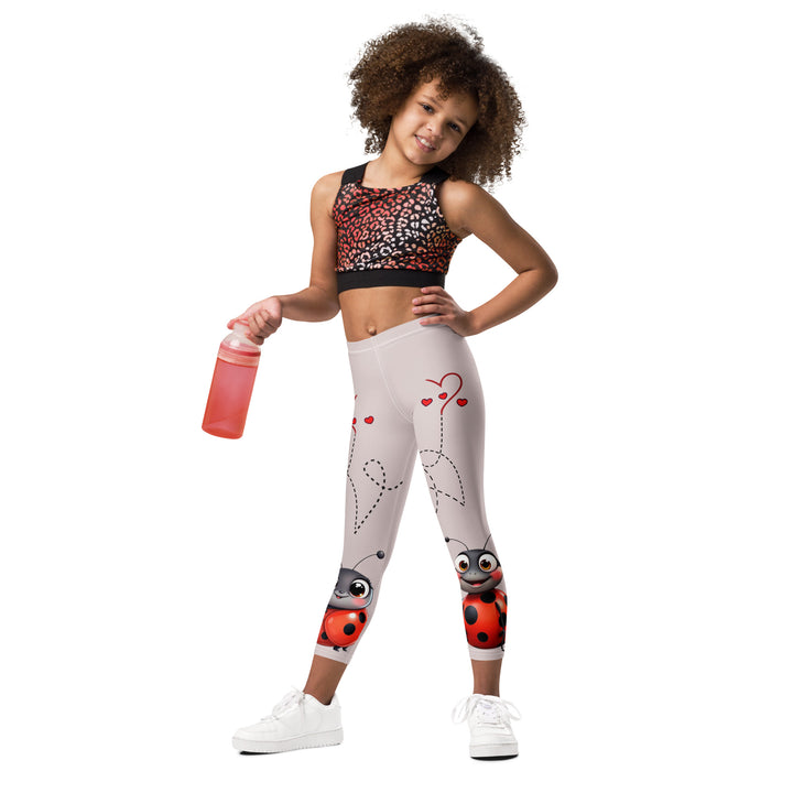 Lovebugs Girls Leggings, sand-colored background with a female ladybug with big eyes at the bottom of one leg and a male ladybug looking delighted on the other leg, with black swirling lines above them forming hearts on both sides, relaxed front pose on model