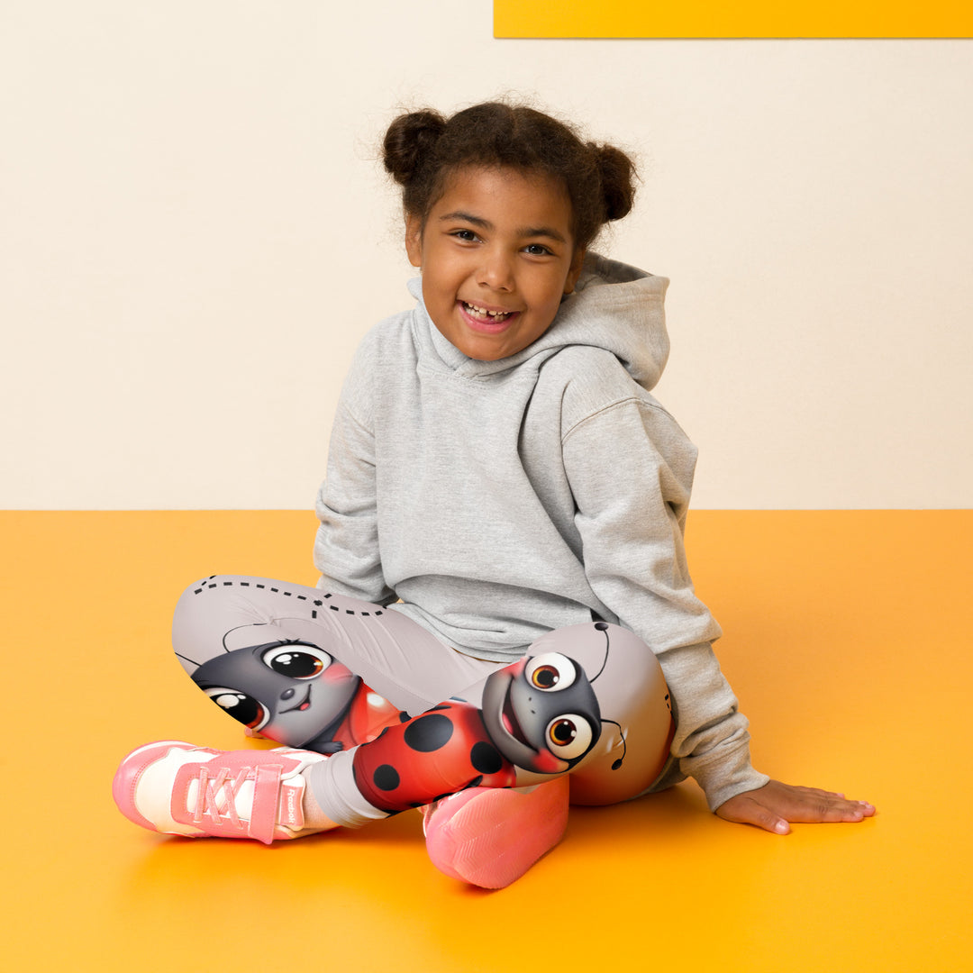  Lovebugs Girls Leggings, sand-colored background with a female ladybug with big eyes at the bottom of one leg and a male ladybug looking delighted on the other leg, with black swirling lines above them forming hearts on both sides, seated pose on model