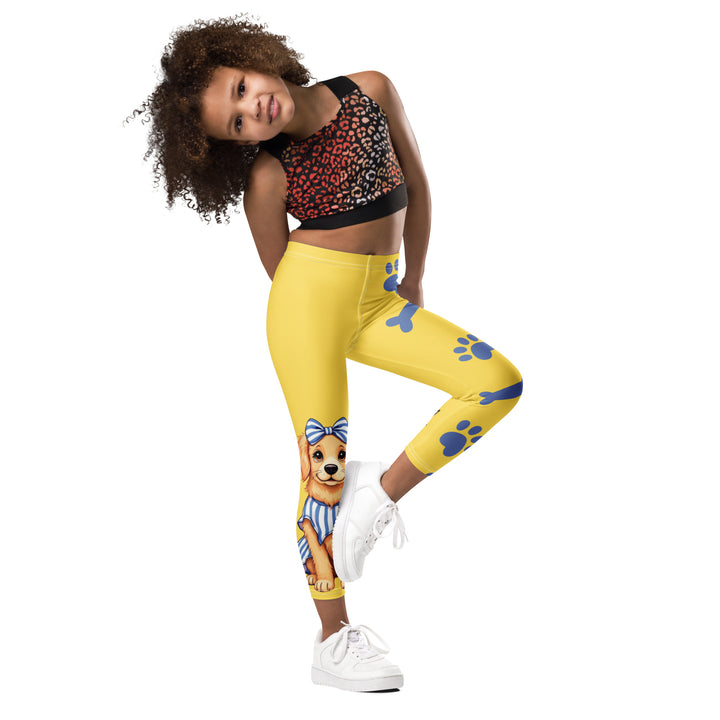 Puppy & Bones Girls Leggings, yellow background with a golden retriever puppy wearing a blue and white striped outfit and a matching blue and white bow on the head on one leg, and blue paw prints and blue bones running down the other leg, side view with one leg lifted and bent on model