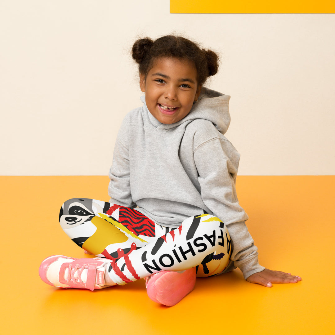 Raccoon Bandit Glow Girls Leggings, white base with splash-like spots in red, purple, and yellow, gray raccoon head, and wavy black stripes, seated pose on model-with a visible 'Leona Fashion' text on the design