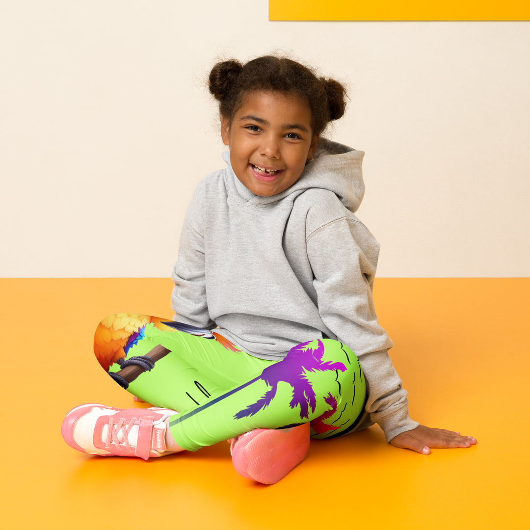 Sunset Parrot Girls Leggings, neon green background with an orange, blue, and purple parrot standing on a branch on one leg, and an orange sunset with flying birds and a purple palm tree on the other leg, seated pose on model