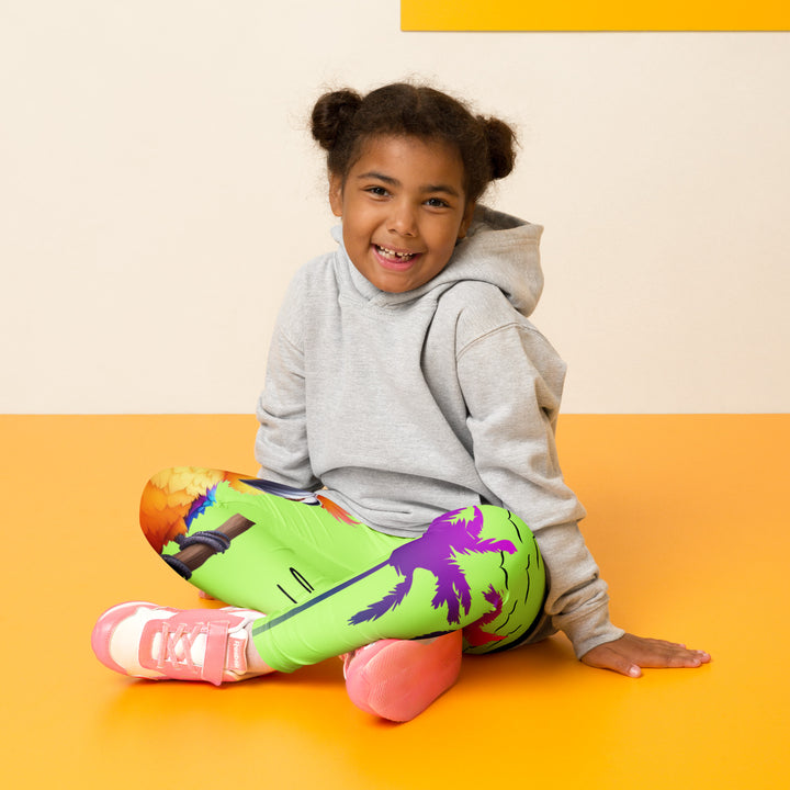Sunset Parrot Girls Leggings, neon green background with an orange, blue, and purple parrot standing on a branch on one leg, and an orange sunset with flying birds and a purple palm tree on the other leg, seated pose on model