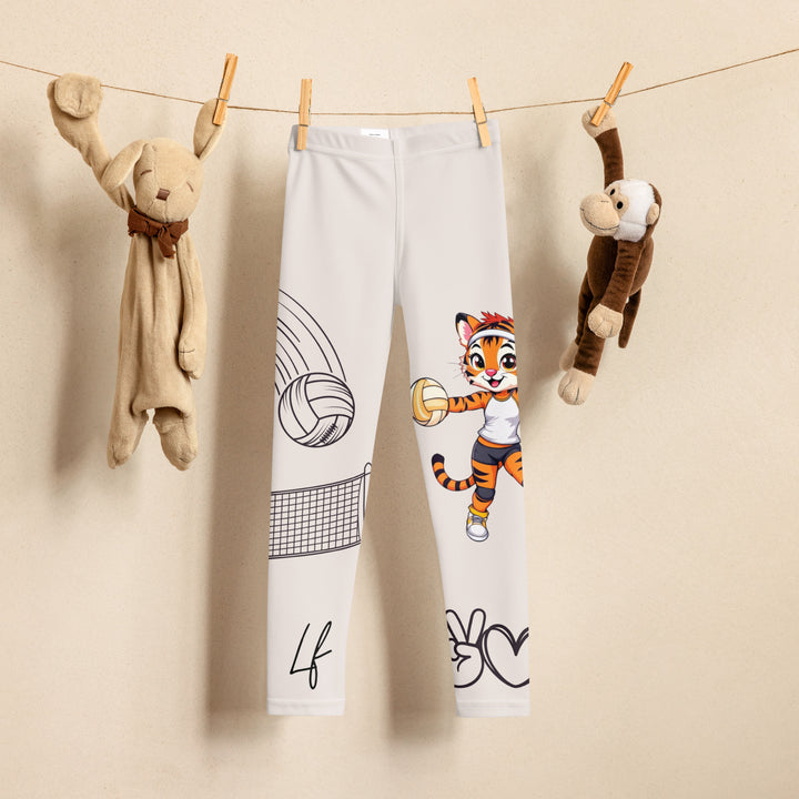  Volley Queen Tiger Girls Leggings, beige background with one leg featuring a sporty dressed tiger holding a volleyball, and the other leg with a volleyball net and a ball leaving a motion trail, front view on hanger