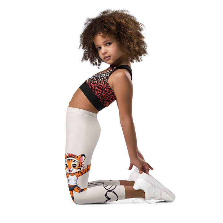  Volley Queen Tiger Girls Leggings, beige background with one leg featuring a sporty dressed tiger holding a volleyball, and the other leg with a volleyball net and a ball leaving a motion trail, left side view kneeling on model