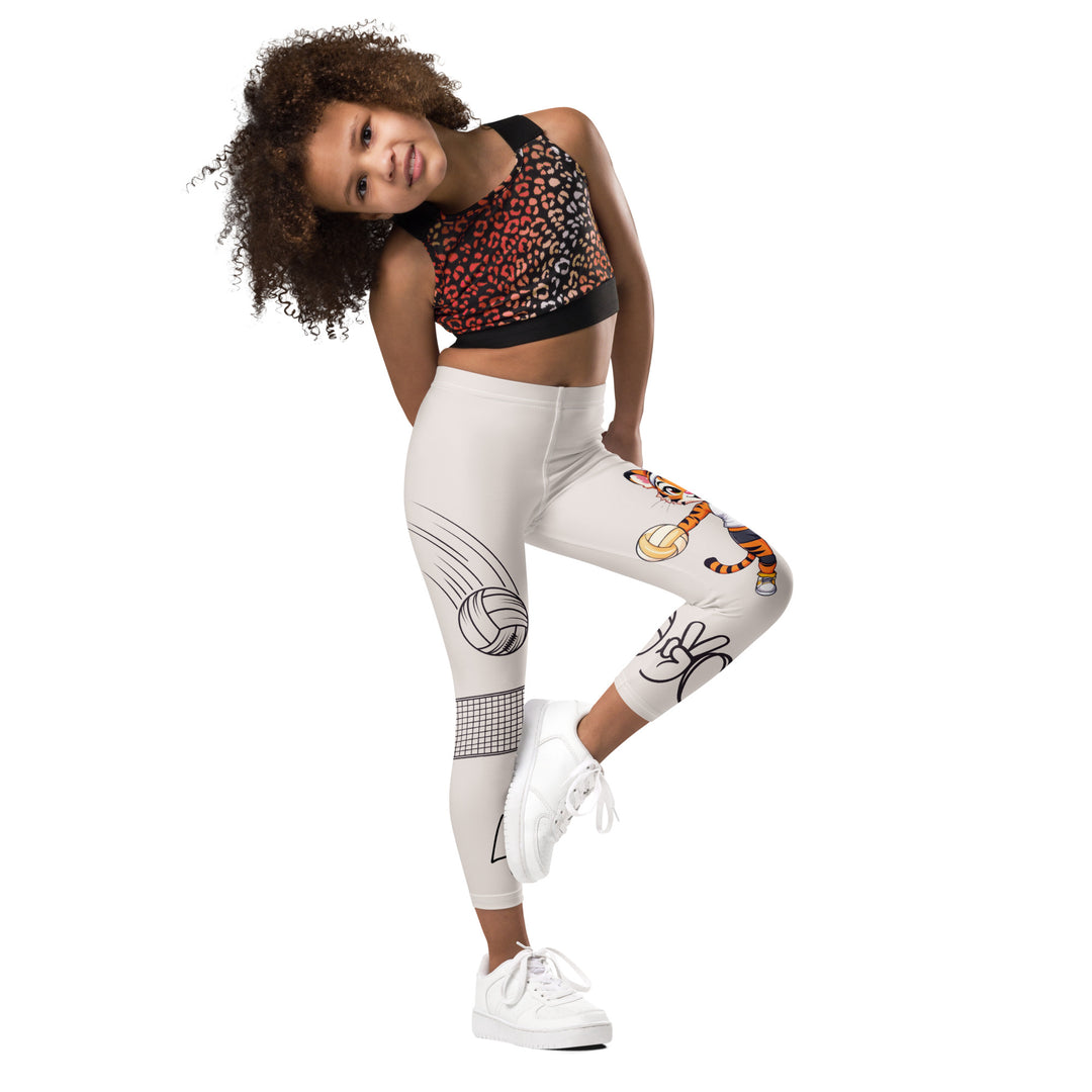  Volley Queen Tiger Girls Leggings, beige background with one leg featuring a sporty dressed tiger holding a volleyball, and the other leg with a volleyball net and a ball leaving a motion trail, side view with one leg lifted and bent on model