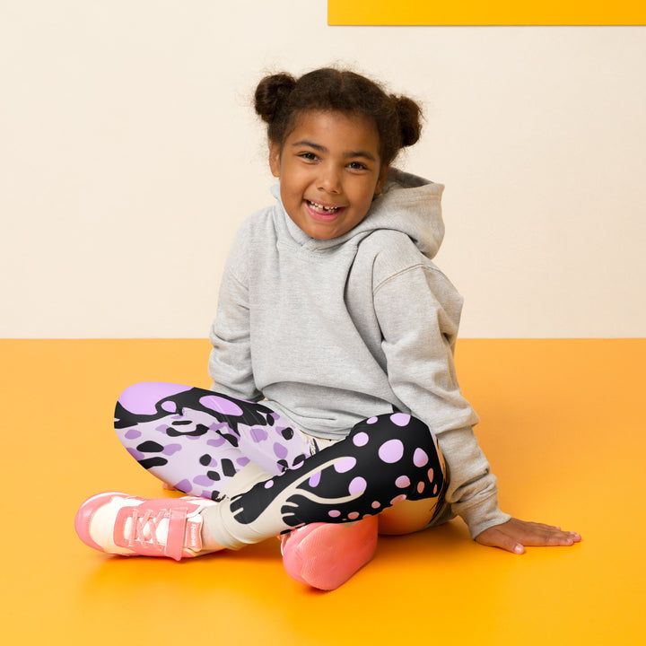 Wild Kitty Paw Purple Girls Leggings, cream background with a black panther featuring soft lavender spots on one leg, and a black and lavender tiger-striped pattern with a large matching paw on the other leg, mixed with cream details, seated pose on model